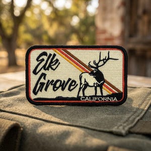 May include: Embroidered patch featuring the words "Elk Grove" in a stylized font, with a silhouette of a deer and the word "California." The patch has a cream-colored background with brown, orange, and yellow stripes.