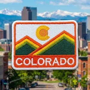 May include: A rectangular embroidered patch featuring a white background with a yellow letter C, green and yellow mountain peaks, and a wide orange base with the word COLORADO in white capital letters, all bordered by an orange frame.