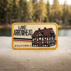 May include: A rectangular embroidered patch with a cream background and a yellow border. It features a black illustration of a house, the text "LAKE ARROWHEAD" in black, "CALIFORNIA" in red, and horizontal red, black, and yellow stripes.