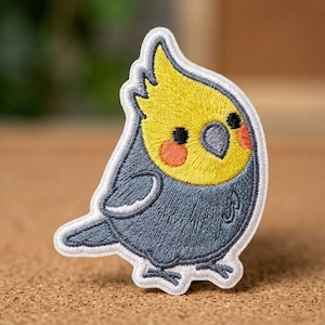 May include: An embroidered patch featuring a cockatiel bird with a bright yellow head, a dark gray body, and orange cheek patches. The bird has a small crest on its head, a gray beak, and black eyes, all outlined with a white stitched border.