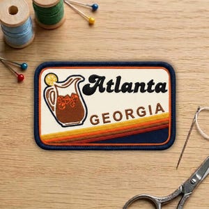May include: Embroidered patch featuring a retro design with the words "Atlanta GEORGIA." The patch includes an illustration of a pitcher of iced tea with a lemon slice. The patch has a navy blue border and a cream-colored background.