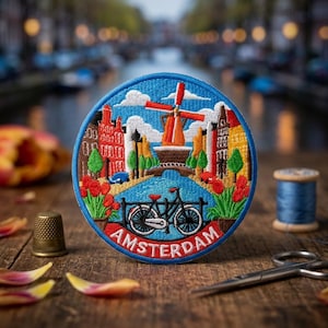 May include: A circular embroidered patch featuring a colorful scene of Amsterdam with a central windmill, canal houses, a bicycle in the foreground, and red tulips. The word AMSTERDAM is stitched in white capital letters along the bottom edge.