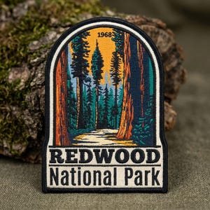 May include: An embroidered patch featuring a forest scene with tall trees, a path, and a yellow sky. The text 1968 appears at the top, while the words REDWOOD National Park are embroidered in black on a cream background at the bottom of the patch.