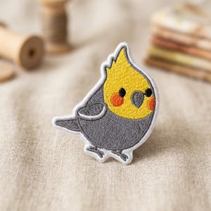 May include: An embroidered patch featuring a stylized cockatiel bird. The bird has a bright yellow head with orange cheek patches, a dark gray body, and a small gray beak. The design is outlined in white thread, creating a clean, defined edge.