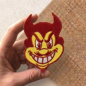 May include: Embroidered patch featuring a cartoon devil with a red head and horns, yellow face, and white teeth. The patch has a smiling expression and is likely for clothing or accessories.