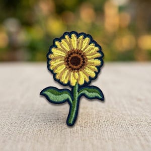May include: An embroidered patch featuring a sunflower with bright yellow petals, a dark brown center, and a green stem with two leaves. The entire design is outlined in a dark navy blue thread, creating a distinct border for the floral motif.