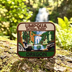 May include: A square embroidered patch featuring a landscape of Umpqua National Forest in Oregon. The design includes a waterfall, green trees, and brown rock formations. The text UMPQUA NATIONAL FOREST is at the top and OREGON is at the bottom.
