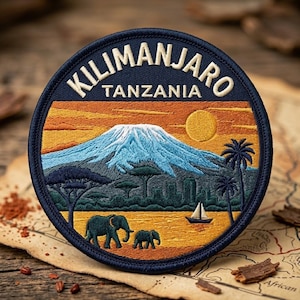 May include: A circular embroidered patch featuring a landscape of Mount Kilimanjaro in Tanzania. The design includes a snow-capped mountain, a yellow sun, palm trees, a sailboat on water, and two elephants in shades of blue, orange, and yellow.