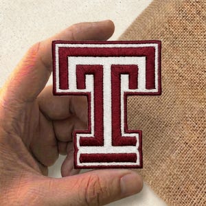 May include: Embroidered patch featuring a maroon and white letter "T". The letter is in a bold, block style with a white border and interior details. The patch is held against a neutral background.