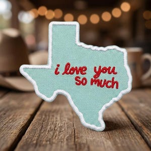 May include: A light blue embroidered patch in the shape of the state of Texas with a white border. The text "i love you so much" is stitched in red thread across the center of the patch, featuring a cursive-style font for the lettering.