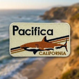 May include: A rectangular embroidered patch with a cream-colored background. It features the black text 'Pacifica' at the top, a brown shark graphic in the center with three horizontal stripes behind it, and the word 'CALIFORNIA' in brown at the bottom.