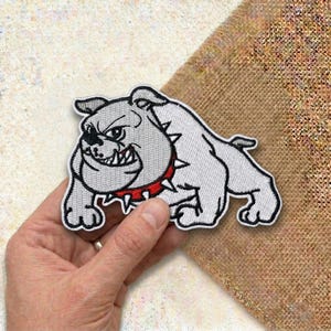 May include: Embroidered patch featuring a gray bulldog with a red spiked collar. The bulldog has an angry expression and is outlined in black. The patch is white with gray shading and is likely for clothing or accessories.