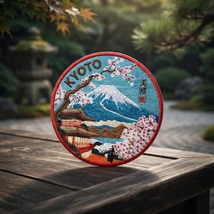 May include: A circular embroidered patch featuring a landscape scene with a snow-capped mountain, a multi-story pagoda, and cherry blossom trees. The text KYOTO is stitched in black at the top, with Japanese characters on the right side.