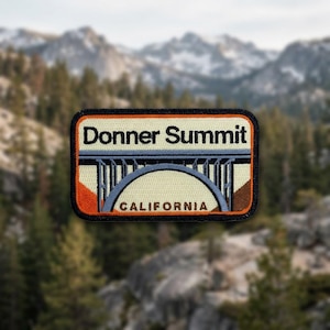 May include: Embroidered patch featuring the words "Donner Summit" above a bridge design, with "CALIFORNIA" below. The patch has an orange border and is set against a blurred backdrop of mountains and trees.