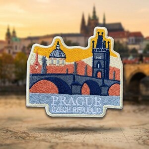 May include: A rectangular embroidered patch featuring a stylized depiction of the Charles Bridge in Prague, Czech Republic. The design includes a dark blue bridge with arches, a tall tower, and a domed building against a yellow and orange sky.