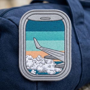 May include: An embroidered patch shaped like an airplane window with a gray frame. The scene inside shows a gray airplane wing extending over white clouds against a horizontal gradient sky of dark blue, light blue, and orange sunset colors.