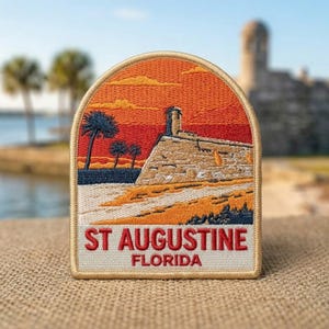 May include: An embroidered patch featuring a sunset scene over a stone fortress and palm trees. The design includes a tan border and the red text ST AUGUSTINE FLORIDA at the bottom. The sky is orange and red, with a tan stone structure and blue water.