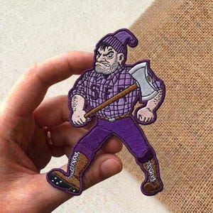 May include: Embroidered patch featuring a cartoon lumberjack in purple attire, holding an axe. The lumberjack wears a purple beanie, plaid shirt with "SF" text, and boots. The patch is held in a hand.