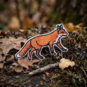May include: An embroidered patch in the shape of a walking fox. The fox features vibrant orange fur with black outlines and white accents on the chest, face, and tail tip. The design is rendered in detailed thread work with a clean white border.