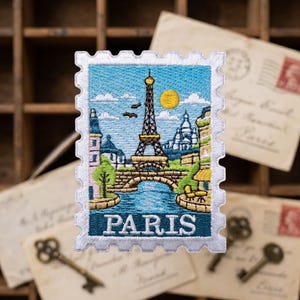 May include: A rectangular embroidered patch shaped like a postage stamp featuring a scene of Paris. The design includes the Eiffel Tower, a stone bridge over a blue river, buildings, a yellow sun, and the word PARIS in white capital letters.
