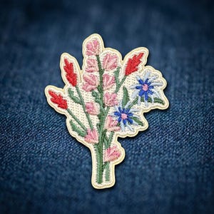 May include: An embroidered patch featuring a bouquet of wildflowers. The design includes pink, red, and blue flowers with green stems and leaves, all stitched onto a cream-colored fabric base with a die-cut edge following the floral shape.