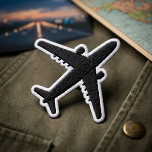 May include: An embroidered patch in the shape of a commercial airplane. The airplane is black with a thick white border outlining the wings, fuselage, and tail. The embroidery features a textured stitch pattern throughout the black surface area.