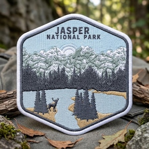 May include: A hexagonal embroidered patch featuring a landscape of Jasper National Park. The design shows a light blue sky, snow-capped mountains, a dark forest, a lake, and a deer standing on the shore. The text reads Jasper National Park.