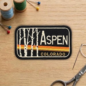 May include: A black embroidered patch with the word "Aspen" in white letters, and "Colorado" in gold. The patch features a design of white birch trees and a retro sunset in shades of orange and yellow.