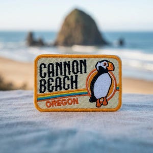 May include: A rectangular embroidered patch with a cream background, featuring the black text CANNON BEACH and orange text OREGON. It includes a colorful horizontal stripe and an illustration of a puffin bird with a black, white, and orange body.