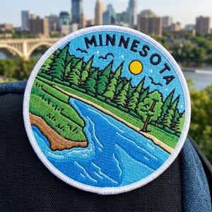 May include: A circular embroidered patch featuring a blue river flowing through a green landscape with pine trees and a yellow sun. The word MINNESOTA is stitched in black letters along the top edge, framed by a white border on a dark fabric.