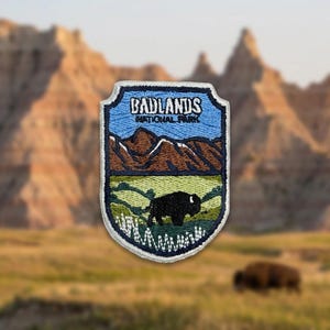 May include: A shield-shaped embroidered patch featuring a landscape of brown mountains under a light blue sky. The text BADLANDS NATIONAL PARK is stitched in white. A black bison stands in a green grassy field in the foreground of the design.