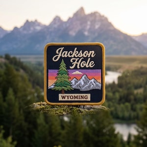 May include: A square embroidered patch with a dark blue border featuring the text Jackson Hole in white script and WYOMING in white block letters. The design includes a green pine tree, grey mountains, and a sunset sky in orange and purple.