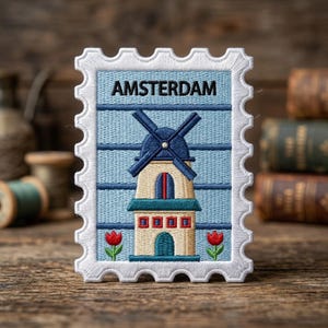 May include: A rectangular embroidered patch shaped like a postage stamp with a white scalloped border. The design features a light blue background with horizontal lines, a central windmill in beige and blue, and two red tulips at the bottom. The text AMSTERDAM is embroidered in black at the top.