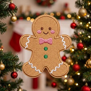 May include: A tan gingerbread man embroidered patch featuring a pink bow tie, two green circular buttons, pink cheeks, and white wavy icing details on the arms and legs. The patch has a stitched border and a smiling face with dark brown eyes.