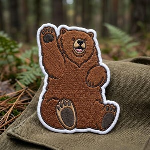 May include: An embroidered patch featuring a brown bear with a white border. The bear is sitting, waving one paw, and has a smiling expression with a visible tongue. The embroidery uses shades of brown for the fur and dark brown for the paw pads.