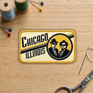 May include: A rectangular patch with the words "Chicago Illinois" in black lettering. The patch features a circular image of two men in suits and hats. The patch has a yellow border and is on a wooden surface.