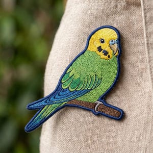 May include: An embroidered patch featuring a budgerigar with a bright yellow head, black cheek spots, and a green body with blue wing feathers. The bird is perched on a brown branch, outlined with a dark blue border against a textured fabric.