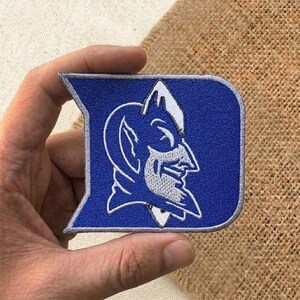 May include: A blue and white embroidered patch featuring the Duke Blue Devils logo. The patch is in the shape of a letter "D" with the team's devil mascot in profile. The patch is held in a hand.