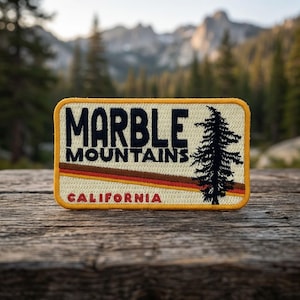 May include: A rectangular embroidered patch with a cream background and a yellow border. It features the dark blue text MARBLE MOUNTAINS, a black pine tree silhouette, two horizontal orange and brown stripes, and the red text CALIFORNIA.