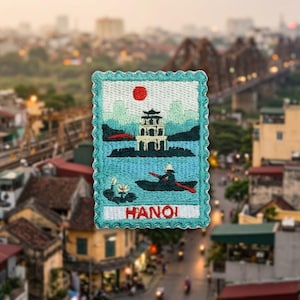 May include: A rectangular embroidered patch with a scalloped teal border. The design features a pagoda on an island, a person in a boat, and a red sun against a light blue sky. The word HANOI is stitched in red at the bottom.