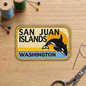 May include: A rectangular patch with a yellow border features the text "SAN JUAN ISLANDS WASHINGTON". A black and white orca whale is depicted in front of a yellow sun. The patch is on a wooden surface with sewing supplies.