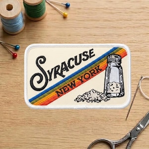 May include: Embroidered patch featuring the text "Syracuse New York" with a salt shaker illustration. The patch has a cream background with a diagonal rainbow stripe. Additional sewing supplies are in the background.