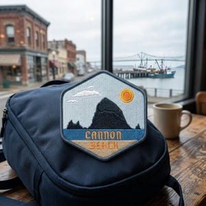 May include: A hexagonal embroidered patch featuring a landscape of Cannon Beach, Oregon. The design shows a dark gray sea stack, a yellow sun, white clouds, blue water, and tan sand. The text CANNON BEACH is stitched in orange on the patch.