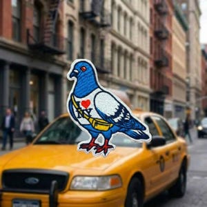 May include: An embroidered patch featuring a blue and white pigeon wearing a yellow crossbody bag. The pigeon has a red heart icon on its chest above the letters NY. The patch has a white border and is shaped like the silhouette of a standing bird.