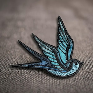 May include: An embroidered swallow patch featuring vibrant turquoise and light blue thread detailing against a bold black stitched outline. The bird is depicted in a dynamic flying pose with wings extended and a long, pointed forked tail.