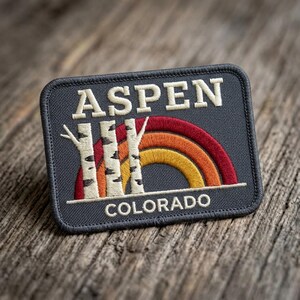 May include: A rectangular embroidered patch with a dark gray background. The text ASPEN appears in white at the top, and COLORADO appears in white at the bottom. Three white birch tree trunks with black markings stand before a red, orange, and yellow rainbow.
