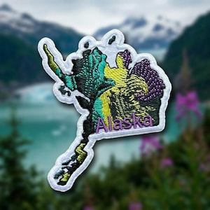 May include: An embroidered patch shaped like the state of Alaska with a white border. The design features a gradient of teal, yellow, and purple thread colors. The word Alaska is stitched in purple thread across the lower section of the patch.