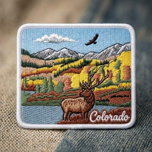 May include: A rectangular embroidered patch featuring a landscape with a brown elk standing near a blue lake, yellow and green trees, snow-capped mountains, a flying bird in a blue sky, and the white cursive text Colorado at the bottom right.