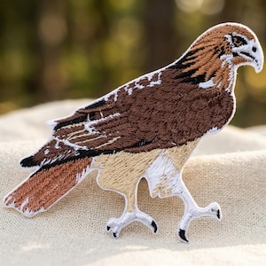 May include: An embroidered iron-on patch featuring a hawk in profile. The bird has brown and tan feathers, a white underbelly, and a white border. The hawk is depicted in a walking stance with its head turned to the right, showcasing detailed stitching.