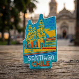 May include: An embroidered patch featuring a stylized architectural design in shades of blue, yellow, and orange. The patch displays the text SANTIAGO CHILE in white and orange lettering at the bottom, with a detailed stitched border and texture.